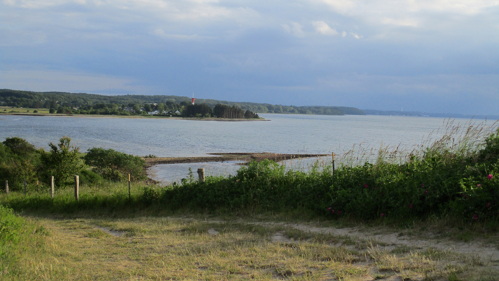 Ein Blick von einem Strandweg, mit Gras und Büschen gesäumt, auf das Meer an einem bewölkten Tag, im Hintergrund sieht man einen kleinen rot-weißen Leuchtturm.